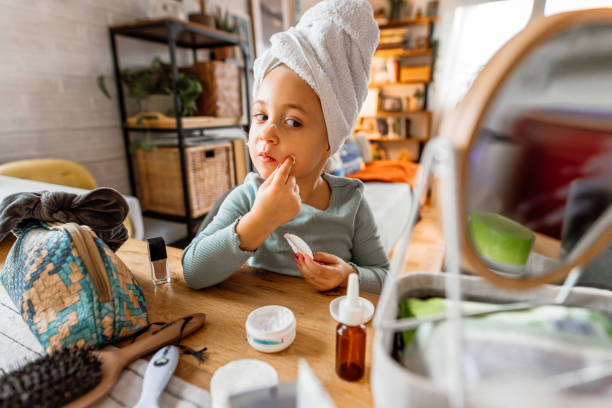 Smiling little girl with towel twisted around head, looking at mirror while applying skin cream on her face.